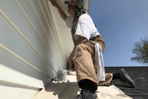 A painter in overalls works on the siding of a house from a roof edge under a clear blue sky.