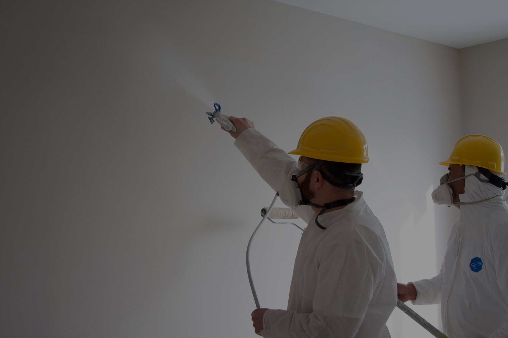 Two construction workers in protective gear spray paint a wall inside a room.